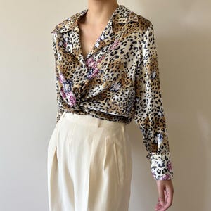 May include: A long-sleeved button-down shirt featuring a brown and black leopard print pattern with scattered dark purple floral accents. The shirt has a pointed collar, buttoned cuffs, and is tucked into a pair of cream-colored high-waisted trousers.