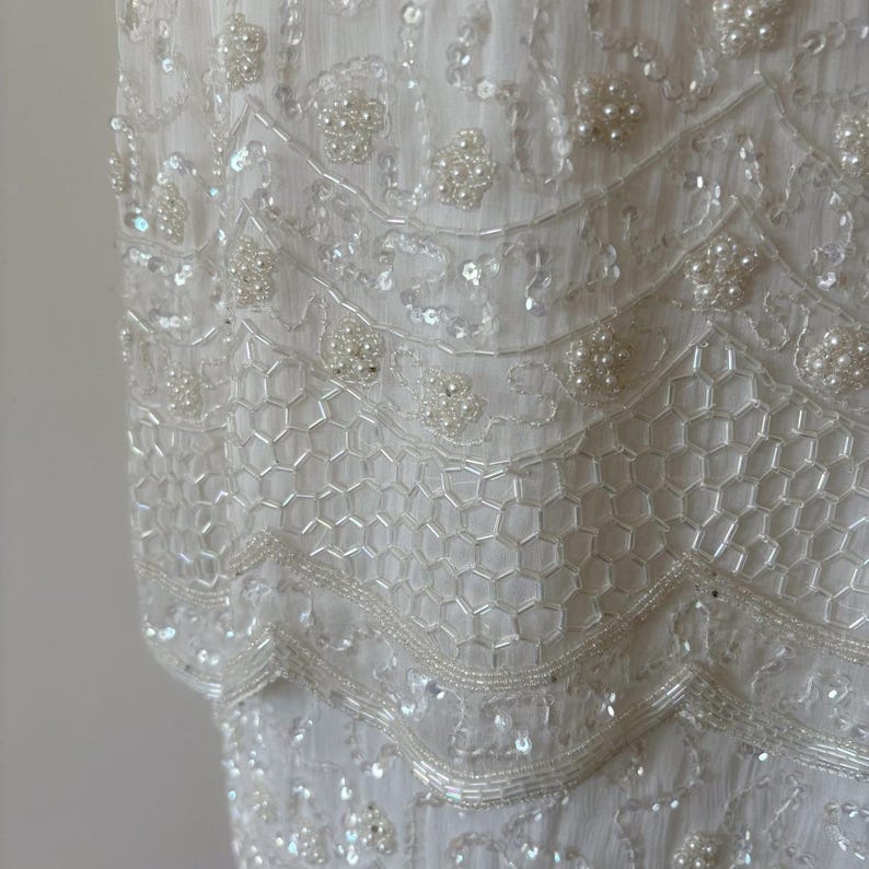 May include: Close-up of a white dress with intricate beadwork and sequin embellishments. The dress features layers of delicate beading, creating a textured, elegant design. The details include pearl-like beads and a honeycomb-like pattern.