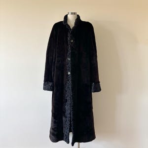 May include: A long, dark brown coat featuring a smooth, plush texture on the main body panels. The garment includes a textured, curly trim along the front placket, collar, and sleeve cuffs, secured by three visible buttons down the center front.