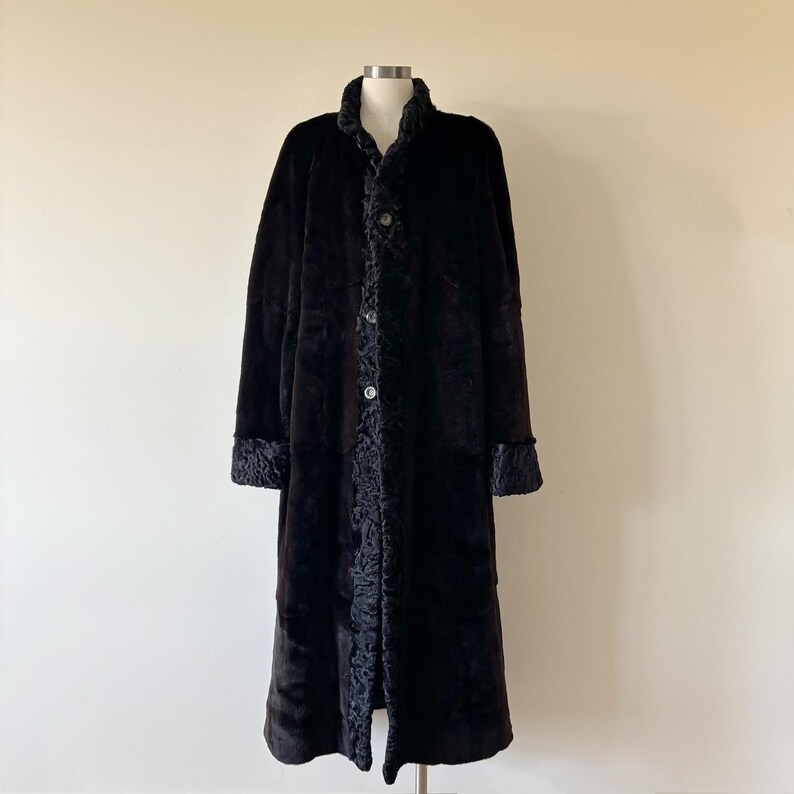 May include: A long, dark brown coat featuring a smooth, plush texture on the main body panels. The garment includes a textured, curly trim along the front placket, collar, and sleeve cuffs, secured by three visible buttons down the center front.