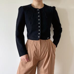Puede incluir: Chaqueta negra de lana con cuello redondo y botones plateados decorativos. La chaqueta tiene mangas largas y largo recortado. La modelo lleva la chaqueta con pantalones de talle alto y plisados color beige. La chaqueta tiene una pequeña etiqueta plateada cerca del dobladillo.