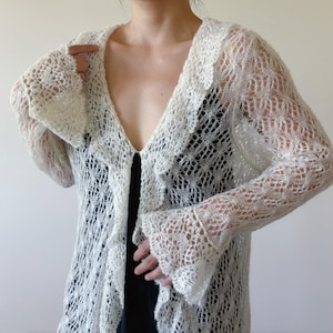 以下が含まれることがあります： 繊細なオープンニットデザインの、クリーム色のオープンフロントカーディガン。長袖セーターは、フリルカラーとフレア袖口が特徴です。軽量の衣服は重ね着に適しており、どんな服装にもエレガントさを加えます。