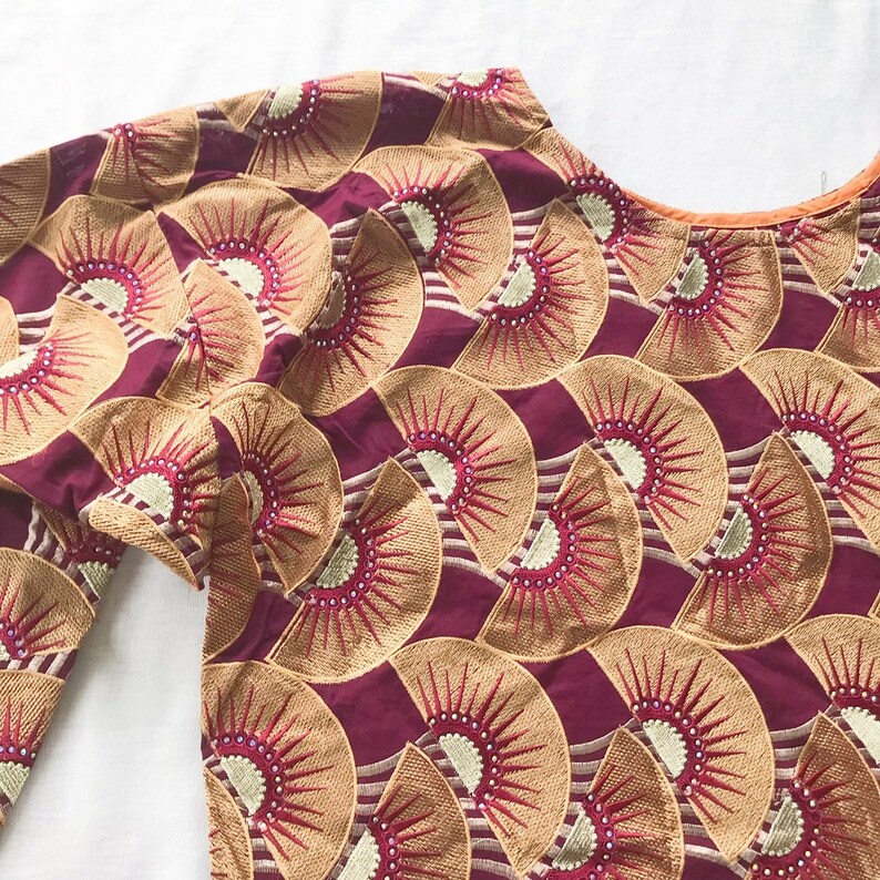 May include: A close-up of a maroon-colored top with long sleeves. The top is adorned with a repeating fan-shaped pattern in gold and red embroidery. The neckline is trimmed with a thin orange border. The fabric has a slight sheen.