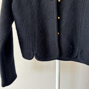 Puede incluir: Primer plano de una chaqueta de lana negra con dobladillo festoneado y botones dorados. La chaqueta tiene una superficie texturizada y un dise&ntilde;o cl&aacute;sico. Las mangas son largas y la chaqueta est&aacute; abotonada.