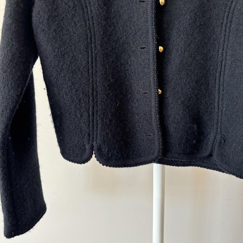 Puede incluir: Primer plano de una chaqueta de lana negra con dobladillo festoneado y botones dorados. La chaqueta tiene una superficie texturizada y un dise&ntilde;o cl&aacute;sico. Las mangas son largas y la chaqueta est&aacute; abotonada.