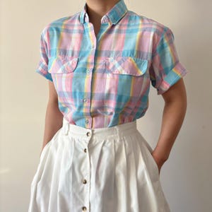 May include: A pastel plaid short-sleeve shirt with two chest pockets and a button-down front. The shirt features a color palette of light blue, pink, and yellow. The shirt is paired with a white skirt.