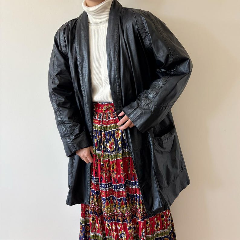 May include: A black leather coat with a relaxed fit, featuring a shawl collar and pockets. The coat is worn over a white turtleneck and a long, pleated skirt with a vibrant floral pattern in red, blue, and green.