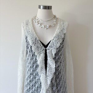 May include: A white, open-front, lace-knit cardigan with a scalloped trim, displayed on a mannequin. A delicate pearl necklace and a black top are visible underneath. The cardigan has long sleeves and a flowing design, perfect for layering.