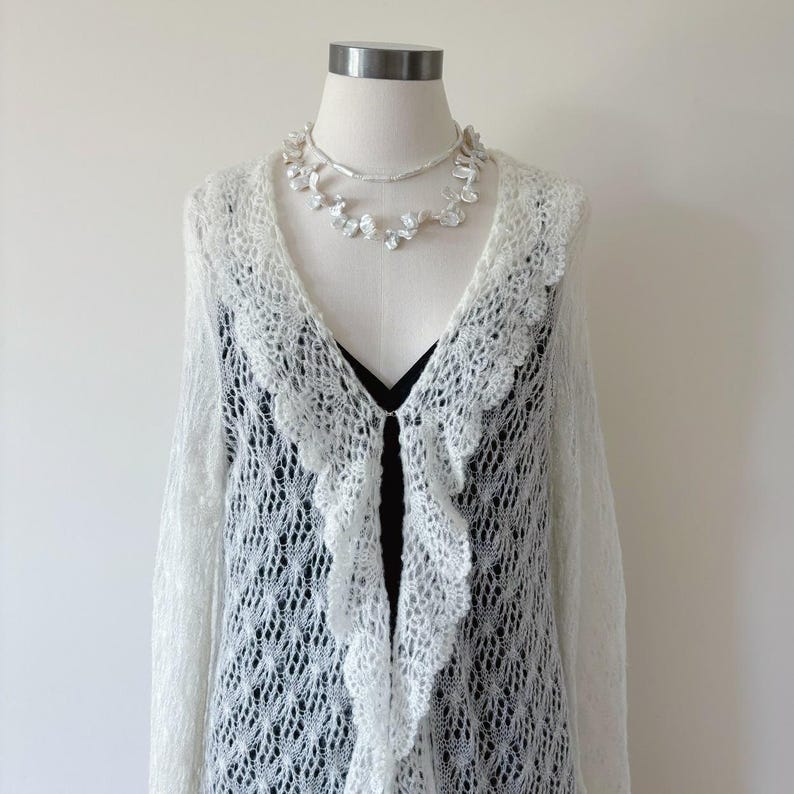 May include: A white, open-front, lace-knit cardigan with a scalloped trim, displayed on a mannequin. A delicate pearl necklace and a black top are visible underneath. The cardigan has long sleeves and a flowing design, perfect for layering.
