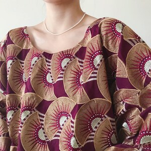 May include: A burgundy-colored top with a square neckline. The fabric is adorned with a repeating pattern of fan-shaped designs in gold and white, creating a textured appearance. The garment has wide sleeves and a loose fit.