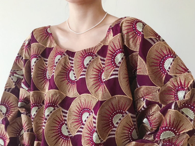 May include: A burgundy-colored top with a square neckline. The fabric is adorned with a repeating pattern of fan-shaped designs in gold and white, creating a textured appearance. The garment has wide sleeves and a loose fit.