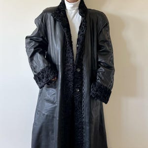 May include: A long black leather coat featuring a textured black faux fur collar and matching cuffs. The coat has a button-front closure, vertical seam detailing, and decorative braided leather trim along the front opening and sleeves.