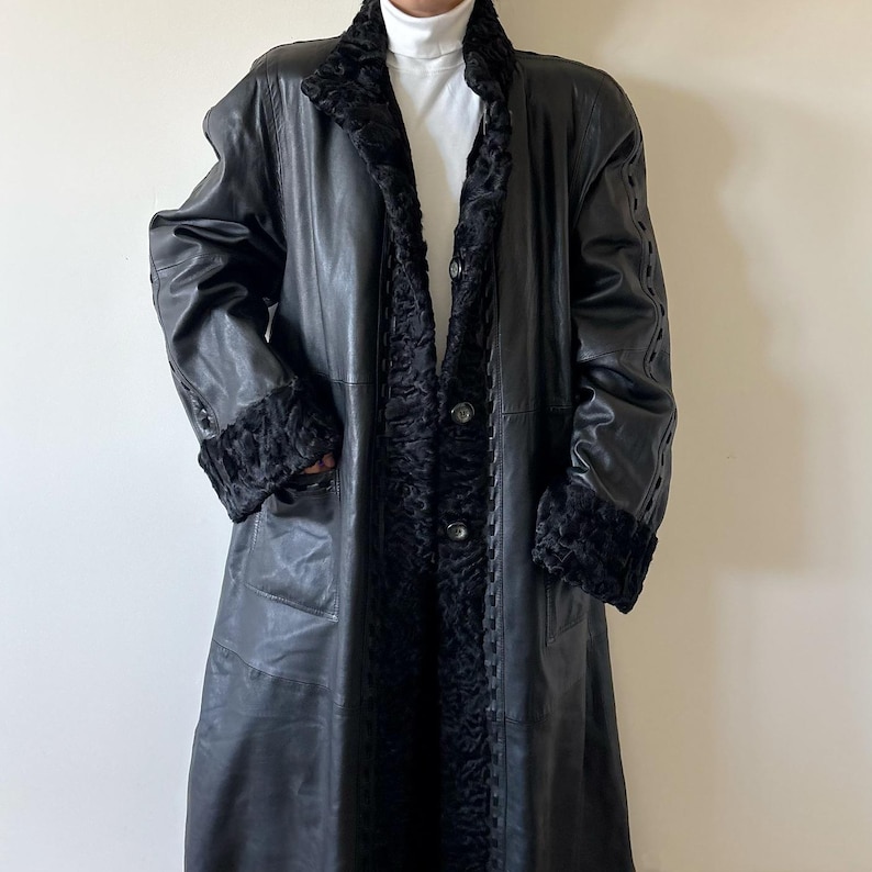 May include: A long black leather coat featuring a textured black faux fur collar and matching cuffs. The coat has a button-front closure, vertical seam detailing, and decorative braided leather trim along the front opening and sleeves.