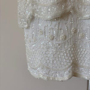 May include: Ivory-colored, beaded dress with a layered design. The dress features intricate beadwork and sequin detailing, with a scalloped hemline. The fabric appears to be a delicate, sheer material.