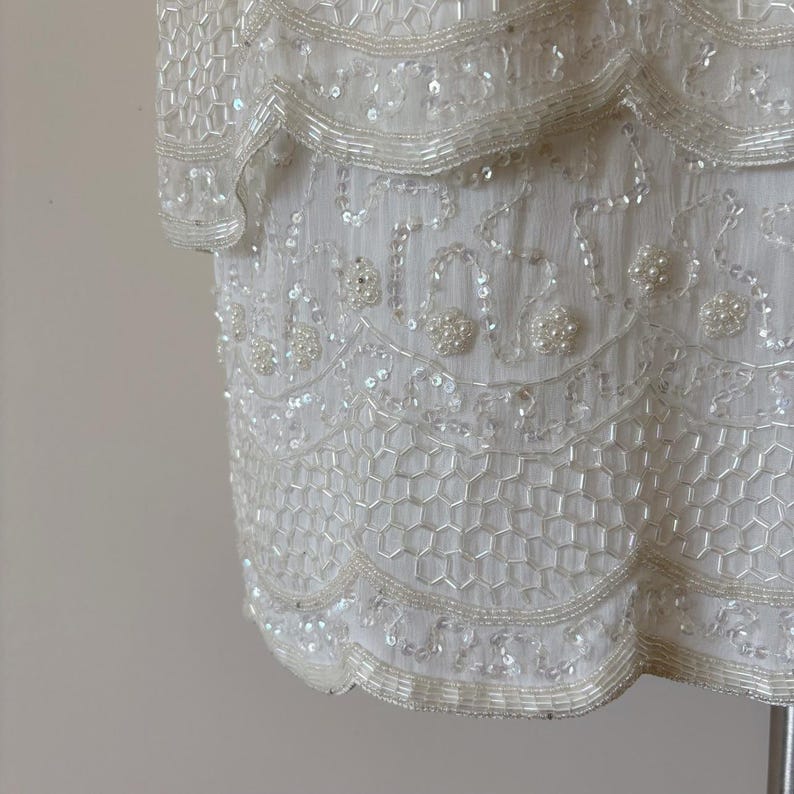 May include: Ivory-colored, beaded dress with a layered design. The dress features intricate beadwork and sequin detailing, with a scalloped hemline. The fabric appears to be a delicate, sheer material.