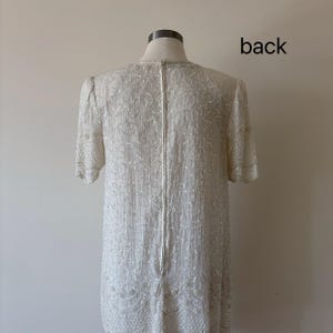 May include: Back view of a cream-colored dress with short sleeves, covered in intricate beadwork. The dress has a straight silhouette and a zipper down the back. The word "back" is written in black on the upper right side.