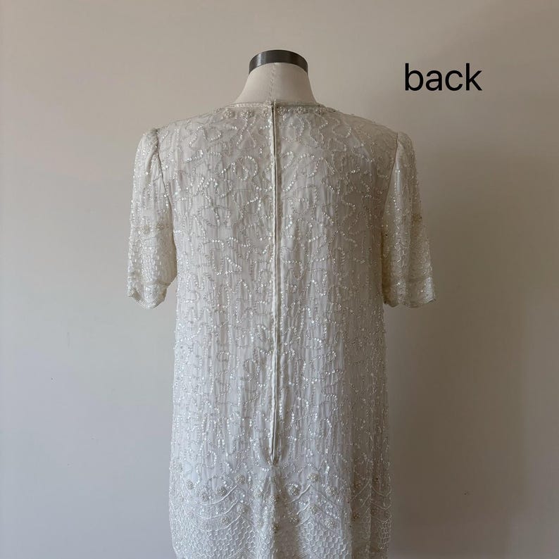 May include: Back view of a cream-colored dress with short sleeves, covered in intricate beadwork. The dress has a straight silhouette and a zipper down the back. The word "back" is written in black on the upper right side.
