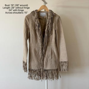 May include: A tan suede jacket with long sleeves and fringe detailing. The jacket has fringe along the collar, front, and sleeves. Bust: 19 inches (38 inches around), Length: 28 inches without fringe, 34 inches with fringe, Across shoulders: 15 inches.