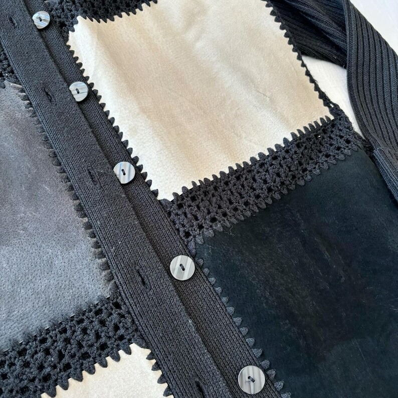 May include: A black cardigan with a patchwork design featuring different textures and colors. The cardigan has a button closure and a crocheted trim.