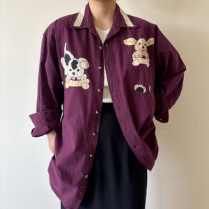 May include: A long-sleeved, button-down shirt in a deep burgundy color. The shirt features appliqued dog designs in cream, black, and tan. The collar has a cream-colored lace trim. The shirt is worn over a white top and black skirt.