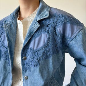 May include: A light blue denim jacket with intricate blue lace detailing on the shoulders and upper chest. The jacket features a classic collar, button closures, and a light wash denim fabric. A white blouse is visible underneath.