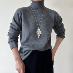 May include: A gray turtleneck sweater paired with black pants. A long silver necklace with a diamond-shaped pendant hangs from the neck. The pendant has a layered design with a circular element in the center.