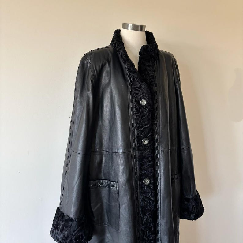May include: A black leather coat featuring a textured black fur-like trim along the collar, front placket, and sleeve cuffs. The coat includes three circular buttons down the center front and decorative woven leather detailing along the seams.