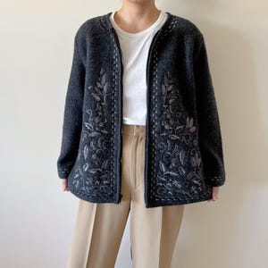 May include: Dark gray zip-up cardigan with embroidered floral design. The cardigan features a round neckline and long sleeves. The floral embroidery is in a lighter shade of gray. The cardigan is worn over a white t-shirt and tan pants.