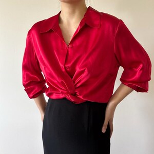 May include: A vibrant red satin blouse with a classic collar and three-quarter sleeves. The blouse features a knotted detail at the waist, paired with a black skirt. The overall look is elegant and stylish.