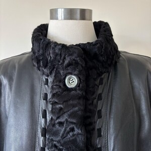 May include: A black leather jacket featuring a textured black faux fur collar and front trim. The garment includes a single silver-toned button closure at the neckline and decorative vertical cut-out detailing along the front placket panels.