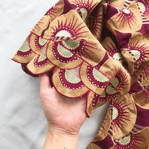 May include: Close-up of a burgundy fabric with intricate embroidery. The design features fan-shaped patterns in gold and red, embellished with small, sparkling rhinestones. The fabric is held in a hand.