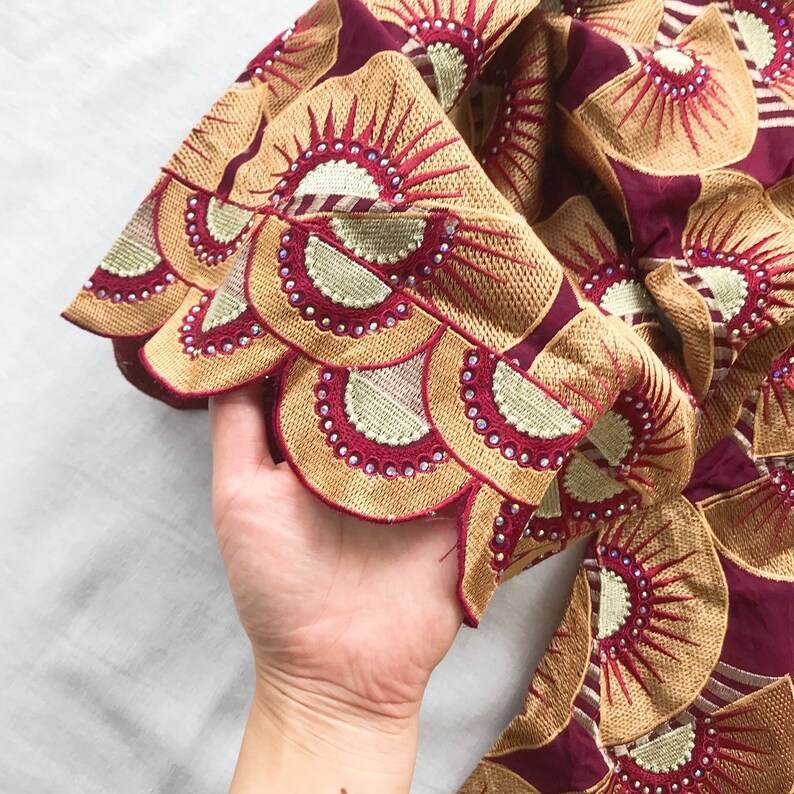 May include: Close-up of a burgundy fabric with intricate embroidery. The design features fan-shaped patterns in gold and red, embellished with small, sparkling rhinestones. The fabric is held in a hand.