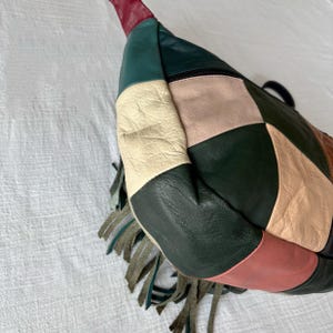 May include: A patchwork leather bag in a variety of colors including green, cream, pink, and red. The bag has a zipper closure and a fringe detail. The bag is a unique accessory.