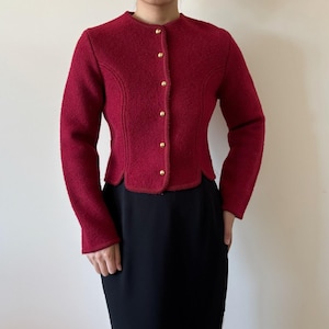 Puede incluir: Chaqueta de lana roja con cuello redondo y botones dorados. La chaqueta tiene mangas largas y un corte entallado. El dobladillo inferior tiene un borde festoneado. La chaqueta se combina con una falda negra.