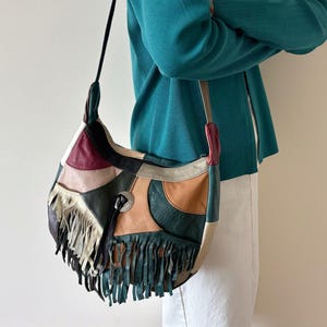 May include: A patchwork shoulder bag with a variety of colors including green, tan, burgundy, and cream. The bag features a long black strap and a fringe detail along the bottom edge. A silver-colored metal accent is visible on the front.