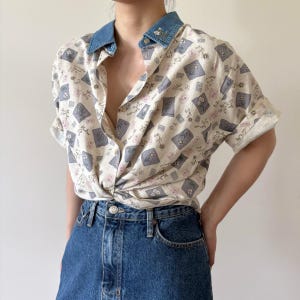 May include: A short-sleeved button-down shirt featuring a cream base with a repeating pattern of gray and pink floral squares. The shirt has a blue denim collar with small floral embroidery and is tucked into dark blue denim jeans.