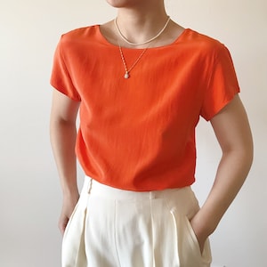 May include: A bright orange short-sleeved blouse with a scoop neck and a simple pearl necklace.