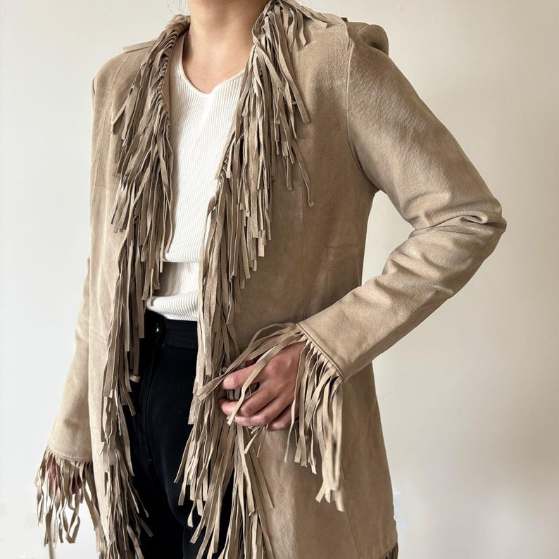 May include: A tan suede jacket with long fringe detailing along the collar, front, and sleeves. The jacket has a long, open design and is worn over a white ribbed top and black pants. The jacket's fringe adds a bohemian flair.