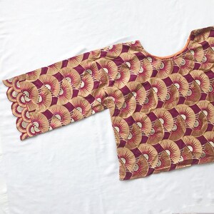 May include: A maroon and gold patterned top with a scalloped sleeve. The fabric features a repeating fan-like design in gold and maroon. The neckline is a rounded scoop shape. The top is displayed on a white surface.