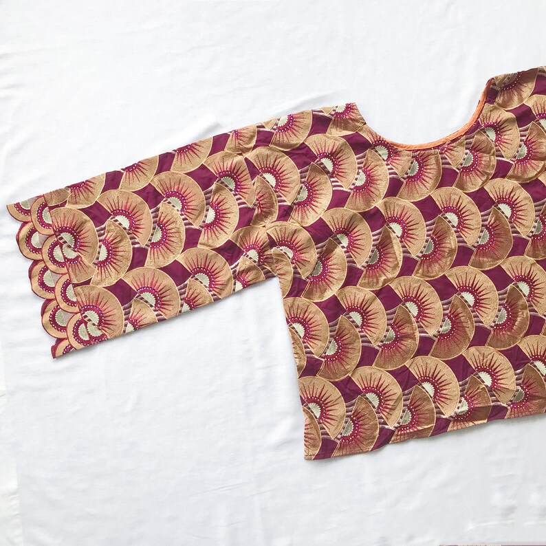 May include: A maroon and gold patterned top with a scalloped sleeve. The fabric features a repeating fan-like design in gold and maroon. The neckline is a rounded scoop shape. The top is displayed on a white surface.