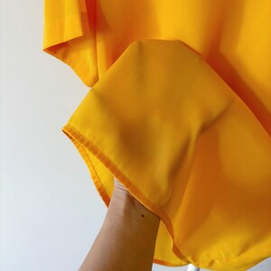Puede incluir: Una camisa de color amarillo brillante con una textura suave y sedosa. La camisa est&aacute; doblada y se sostiene en una mano, mostrando el drapeado de la tela.