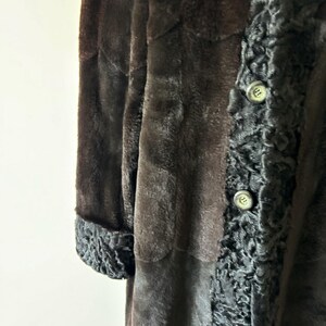 May include: A long coat featuring a smooth dark brown main body and contrasting black textured trim along the front placket and sleeve cuffs. The garment includes two visible round buttons on the front panel with a distinct curly pile texture.