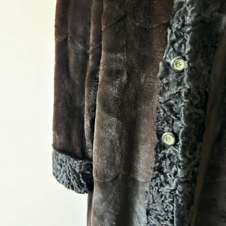 May include: A long coat featuring a smooth dark brown main body and contrasting black textured trim along the front placket and sleeve cuffs. The garment includes two visible round buttons on the front panel with a distinct curly pile texture.