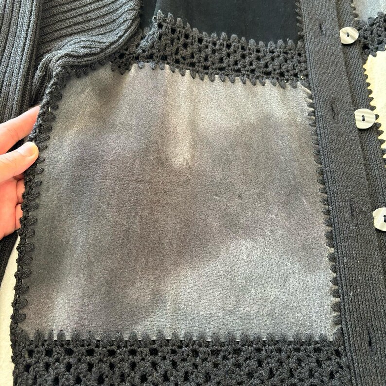 May include: Black cardigan sweater with a crocheted trim and a suede patch on the front. The cardigan has a button closure.