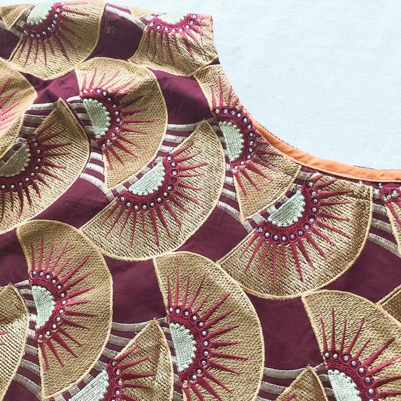 May include: Close-up of a maroon fabric with a repeating fan-shaped embroidered pattern in gold and red. The design features a series of radiating lines and small silver beads. The neckline is trimmed with orange fabric.