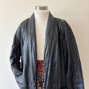 May include: A black leather jacket with a shawl collar is displayed on a mannequin. The jacket is worn over a white turtleneck sweater and a patterned skirt with red, blue, and yellow floral designs. The jacket has a relaxed fit.