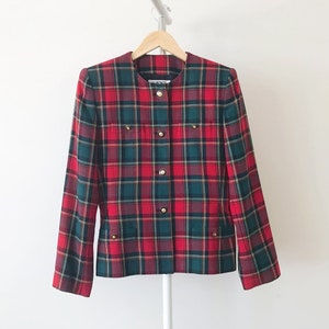 Puede incluir: Blazer de cuadros rojos, verdes y amarillos con botones dorados. La blazer tiene un cuello redondo y mangas largas.