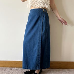 May include: A long, dark blue denim wrap skirt featuring a high-waisted design with a metallic buckle closure at the side. The skirt has a straight silhouette that extends to the ankles, constructed from a durable cotton-based fabric with visible seams.