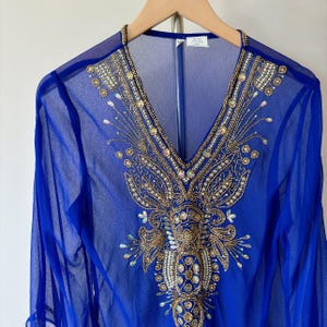 May include: A royal blue sheer top with a gold and pearl beaded design. The top has a V-neck and long sleeves.