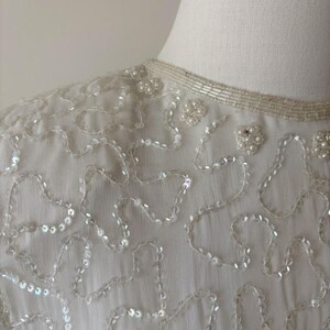 May include: Close-up of a white, vintage top with intricate sequin and bead detailing. The neckline is adorned with delicate beading, and the fabric features flowing, organic patterns of sequins. The overall design is elegant and suitable for formal occasions.